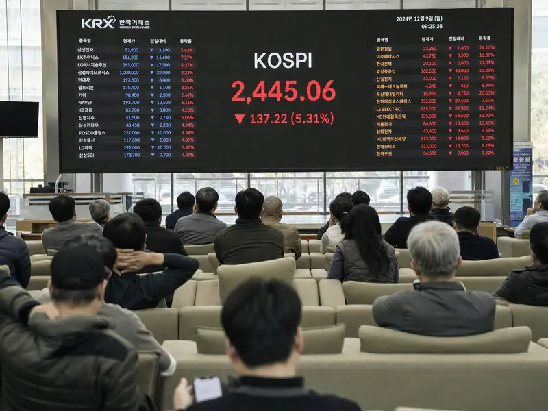 A wide interior shot of retail investors seated in a Seoul securities brokerage watching a large electronic board displaying tumbling KOSPI figures in red, natu