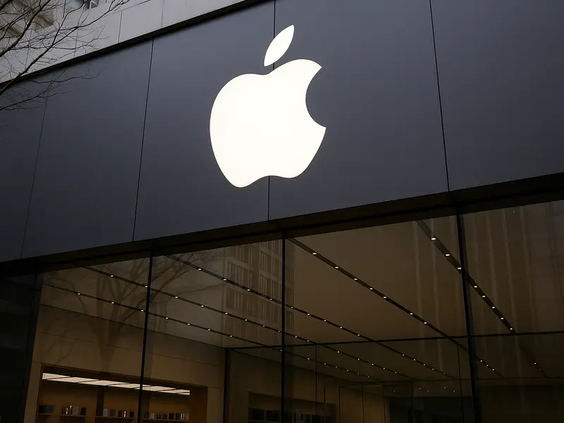 A candid street-level photograph of customers browsing MacBooks and iPads on display through the glass of a busy Apple Store, with the glowing Apple logo above