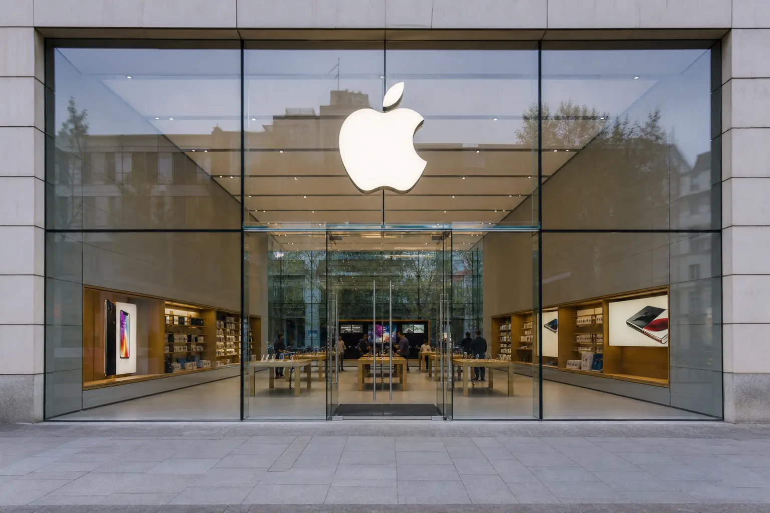 An Apple retail store seen straight-on from the sidewalk in ordinary daylight, its glass frontage and illuminated logo above the entrance.