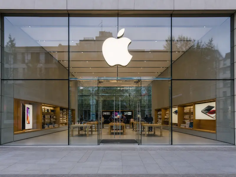 An Apple retail store seen straight-on from the sidewalk in ordinary daylight, its glass frontage and illuminated logo above the entrance.