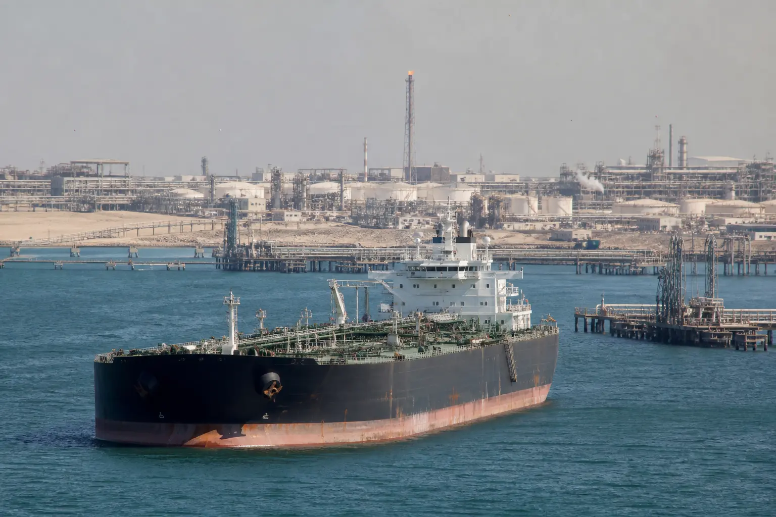 A wide daytime shot of a loaded oil tanker riding low in the water as it transits a Persian Gulf shipping lane, with an oil export terminal's pipework and loadi