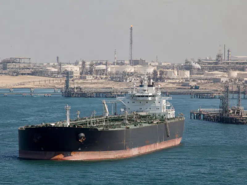 A wide daytime shot of a loaded oil tanker riding low in the water as it transits a Persian Gulf shipping lane, with an oil export terminal's pipework and loadi