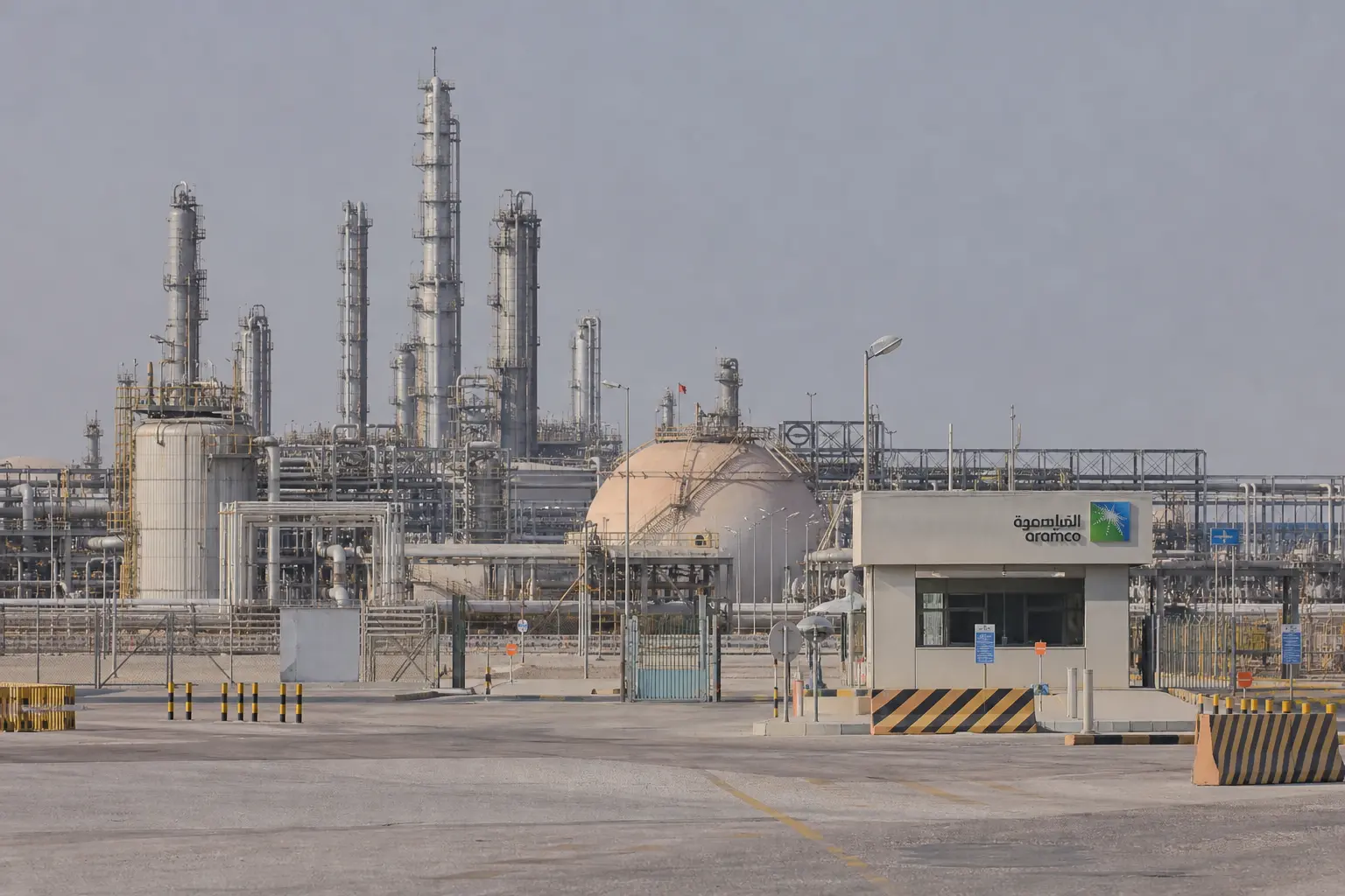 A wide daytime establishing shot of a Saudi Aramco crude oil processing facility with pipelines, storage tanks and distillation towers under a hazy desert sky,