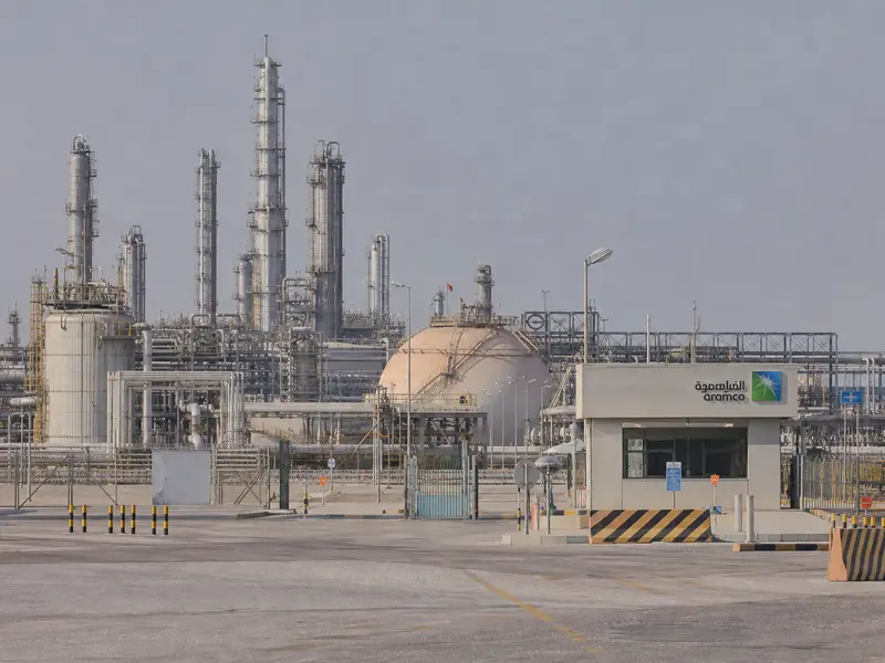 A wide daytime establishing shot of a Saudi Aramco crude oil processing facility with pipelines, storage tanks and distillation towers under a hazy desert sky,