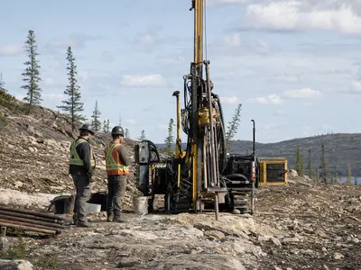 A wide daylight documentary shot of a compact drilling rig operating on a rocky Canadian mineral exploration site, with two technicians in high-visibility vests