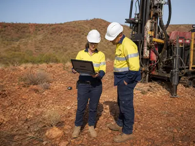 A wide daylight shot of two engineers in hi-vis vests and hard hats standing beside a compact drilling rig at a mineral exploration site, one holding a rugged l