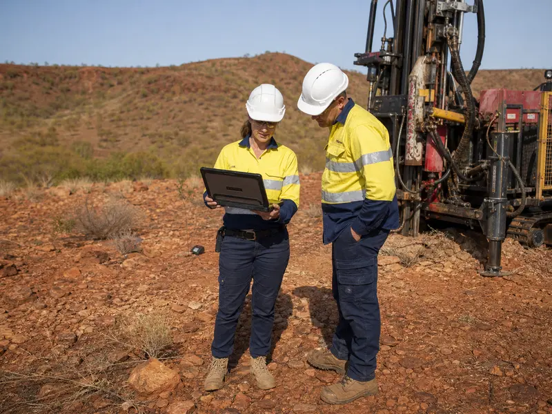 A wide daylight shot of two engineers in hi-vis vests and hard hats standing beside a compact drilling rig at a mineral exploration site, one holding a rugged l