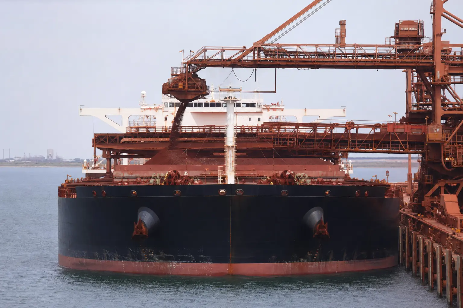 A large bulk iron ore carrier docked head-on at a Pilbara export terminal being loaded with reddish iron ore under flat daylight.