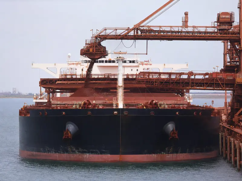 A large bulk iron ore carrier docked head-on at a Pilbara export terminal being loaded with reddish iron ore under flat daylight.