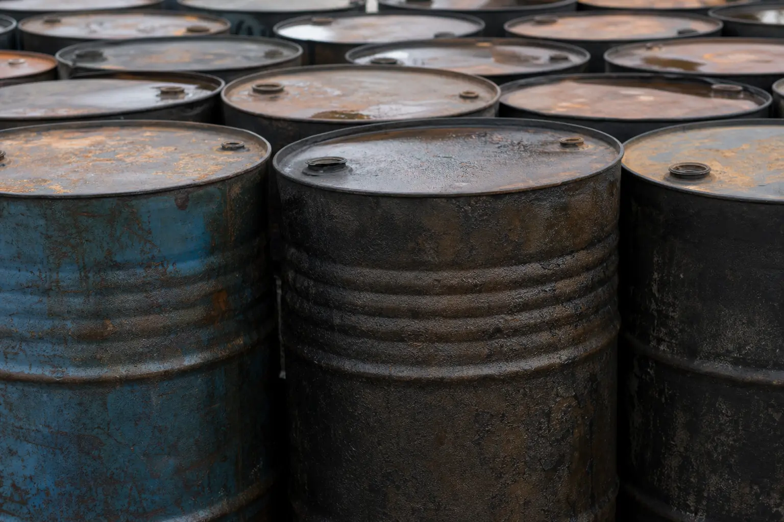 A close-up daylight shot of weathered steel crude oil drums lined up in rows at an industrial storage yard, their rusted lids and ridged sides filling the frame