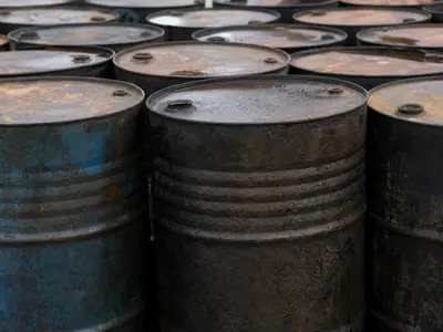 A close-up daylight shot of weathered steel crude oil drums lined up in rows at an industrial storage yard, their rusted lids and ridged sides filling the frame