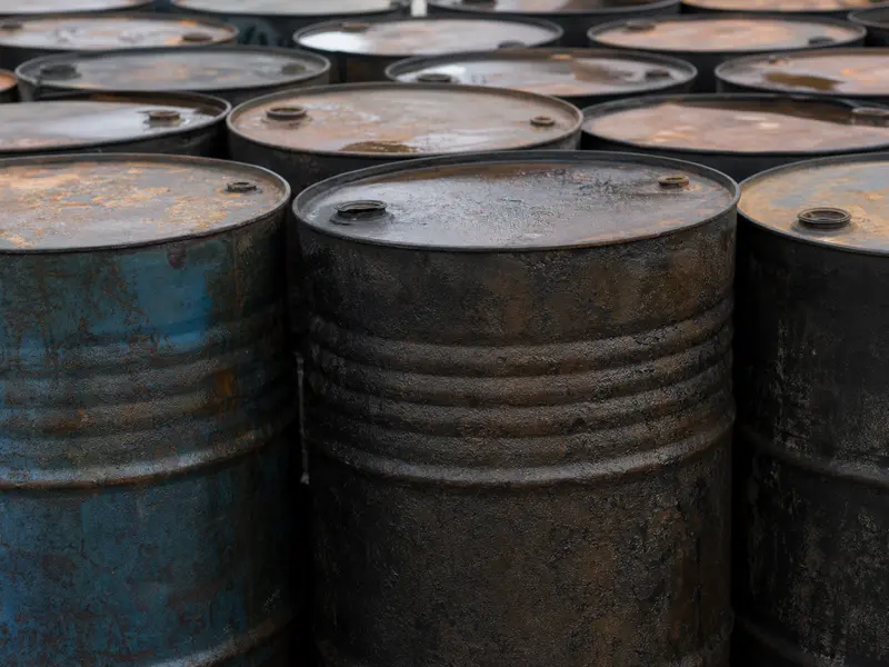 A close-up daylight shot of weathered steel crude oil drums lined up in rows at an industrial storage yard, their rusted lids and ridged sides filling the frame