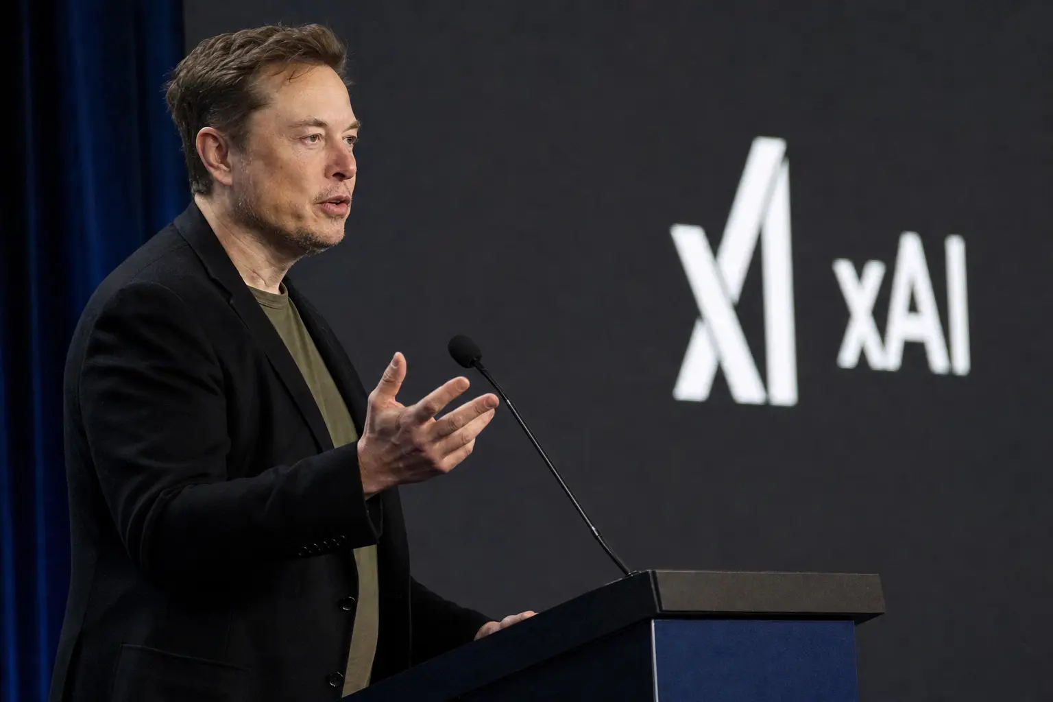 A candid side-view photograph of Elon Musk speaking mid-gesture at a podium during a tech conference, with an xAI logo visible on the backdrop screen behind him