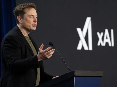 A candid side-view photograph of Elon Musk speaking mid-gesture at a podium during a tech conference, with an xAI logo visible on the backdrop screen behind him