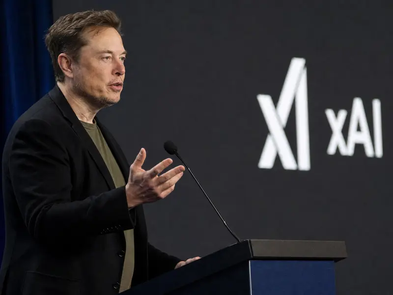 A candid side-view photograph of Elon Musk speaking mid-gesture at a podium during a tech conference, with an xAI logo visible on the backdrop screen behind him
