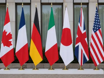 The row of national flags of the G7 member countries standing side by side outside a summit venue, photographed head-on in flat natural daylight.