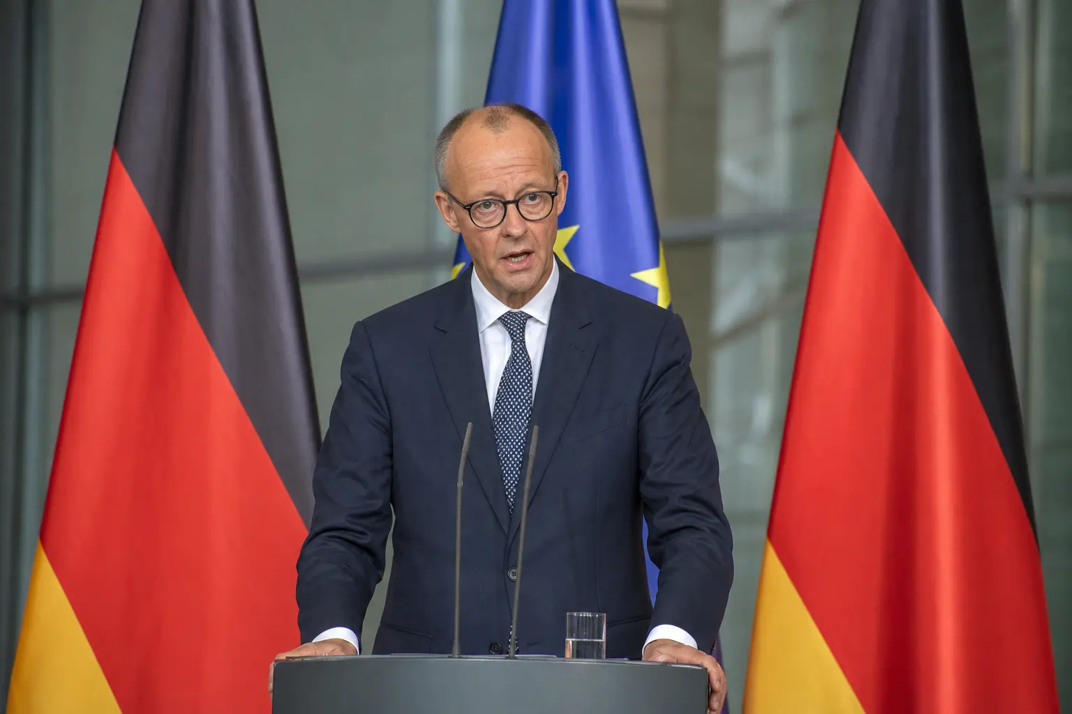 A straight front-on daylight shot of German Chancellor Friedrich Merz speaking at a lectern during a press conference, with German and EU flags standing on pole