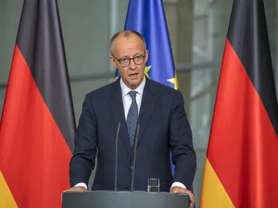A straight front-on daylight shot of German Chancellor Friedrich Merz speaking at a lectern during a press conference, with German and EU flags standing on pole