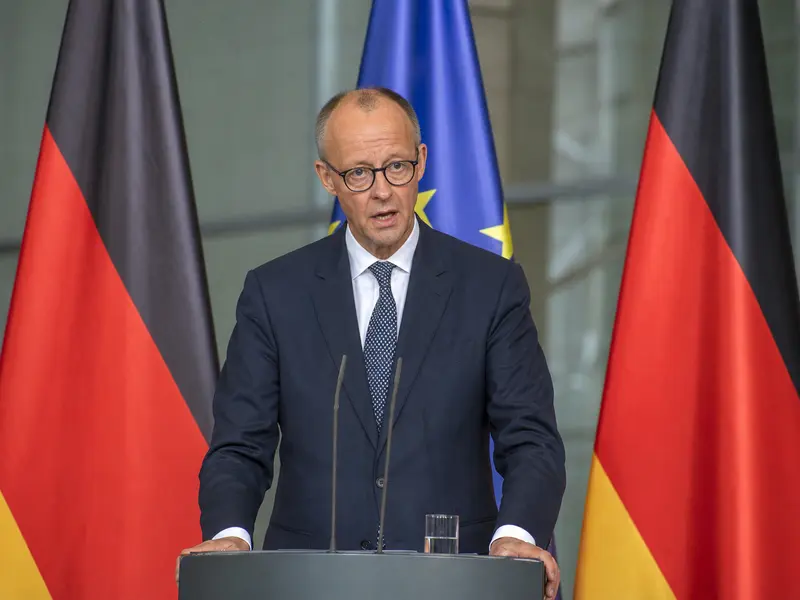 A straight front-on daylight shot of German Chancellor Friedrich Merz speaking at a lectern during a press conference, with German and EU flags standing on pole