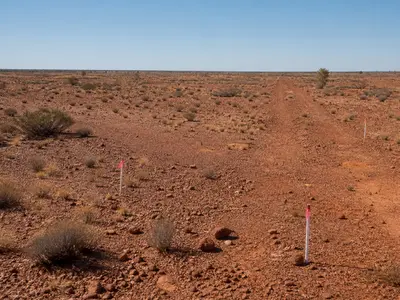 A wide establishing shot of the arid, scrub-covered red-earth terrain of a remote Western Australian lithium project site under bright midday daylight, with a d