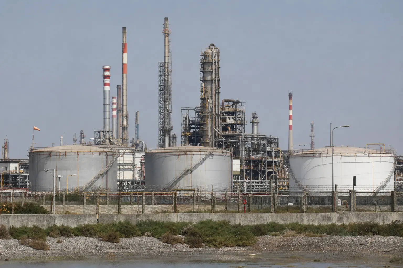 An Indian coastal oil refinery seen head-on in flat daylight, with rows of large crude storage tanks and processing towers behind a perimeter fence.