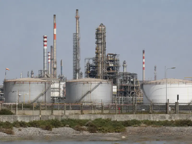 An Indian coastal oil refinery seen head-on in flat daylight, with rows of large crude storage tanks and processing towers behind a perimeter fence.