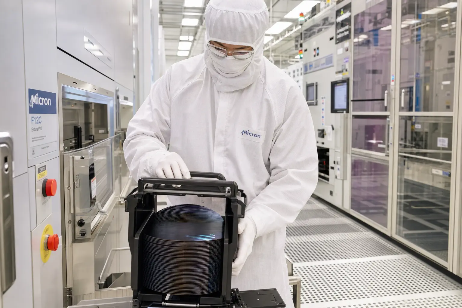 A wide interior shot of a gowned Micron technician in full cleanroom bunny suit handling a silicon wafer cassette inside a brightly lit semiconductor fab, the c