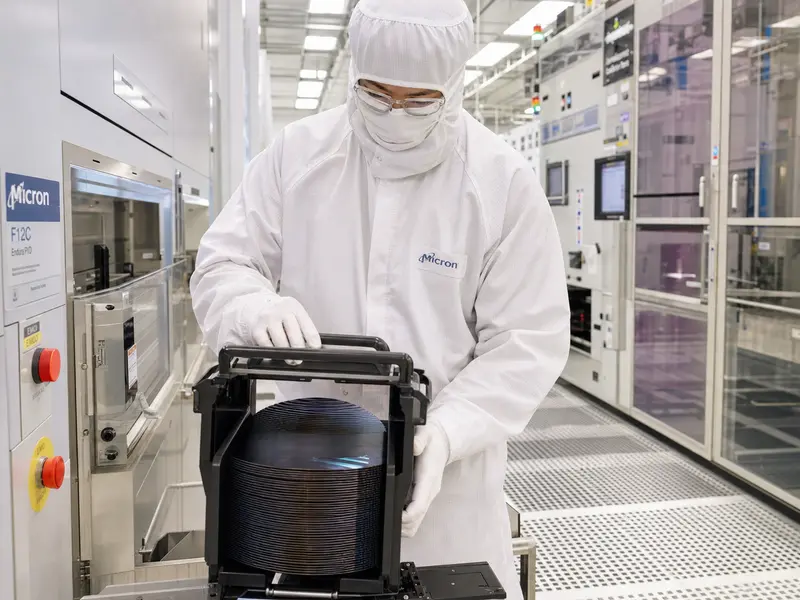 A wide interior shot of a gowned Micron technician in full cleanroom bunny suit handling a silicon wafer cassette inside a brightly lit semiconductor fab, the c