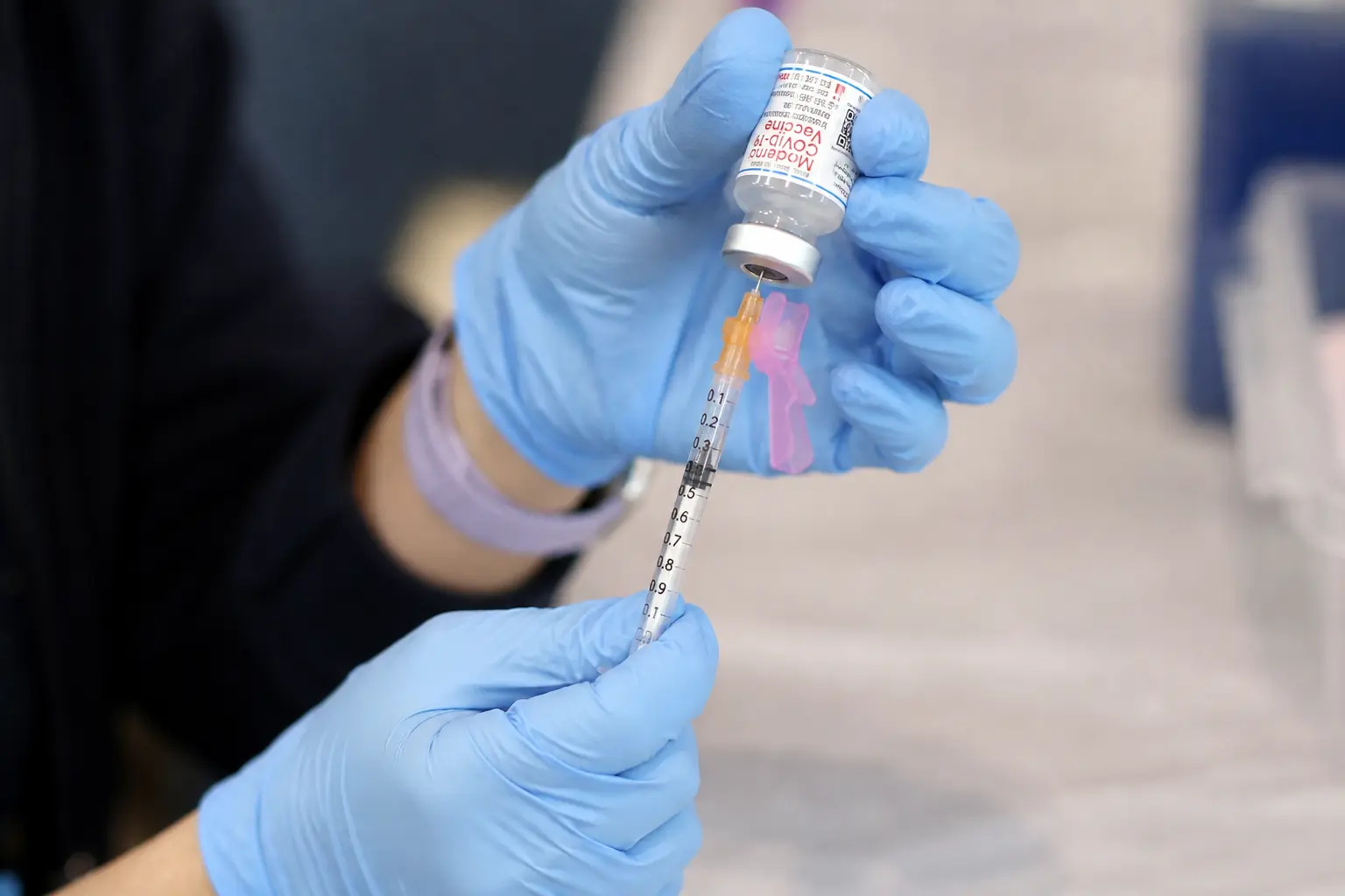 A close, straight-on view of a gloved clinician's hands drawing a Moderna vaccine dose from a small glass vial into a syringe at a vaccination clinic table, the