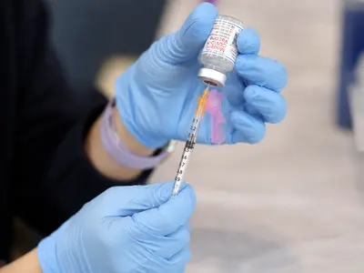 A close, straight-on view of a gloved clinician's hands drawing a Moderna vaccine dose from a small glass vial into a syringe at a vaccination clinic table, the