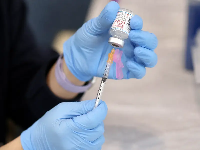 A close, straight-on view of a gloved clinician's hands drawing a Moderna vaccine dose from a small glass vial into a syringe at a vaccination clinic table, the