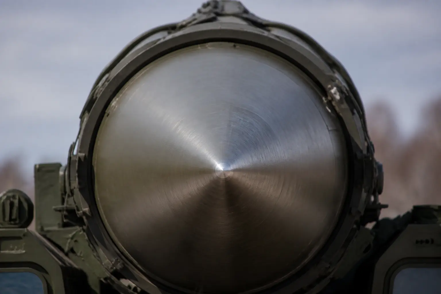A tight close-up of the polished metal nosecone tip of a ballistic missile mounted on its transporter, the heat-resistant alloy surface catching daylight, with