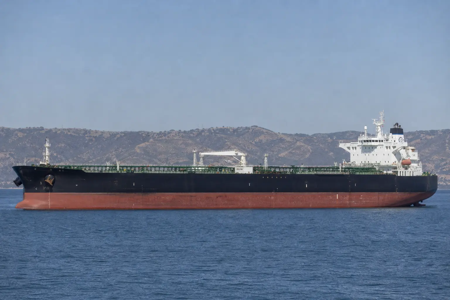 A large crude oil tanker seen broadside underway on calm Gulf waters under flat daylight, low hills of the strait faintly visible on the horizon behind it.