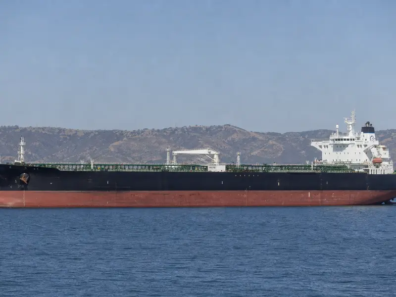 A large crude oil tanker seen broadside underway on calm Gulf waters under flat daylight, low hills of the strait faintly visible on the horizon behind it.