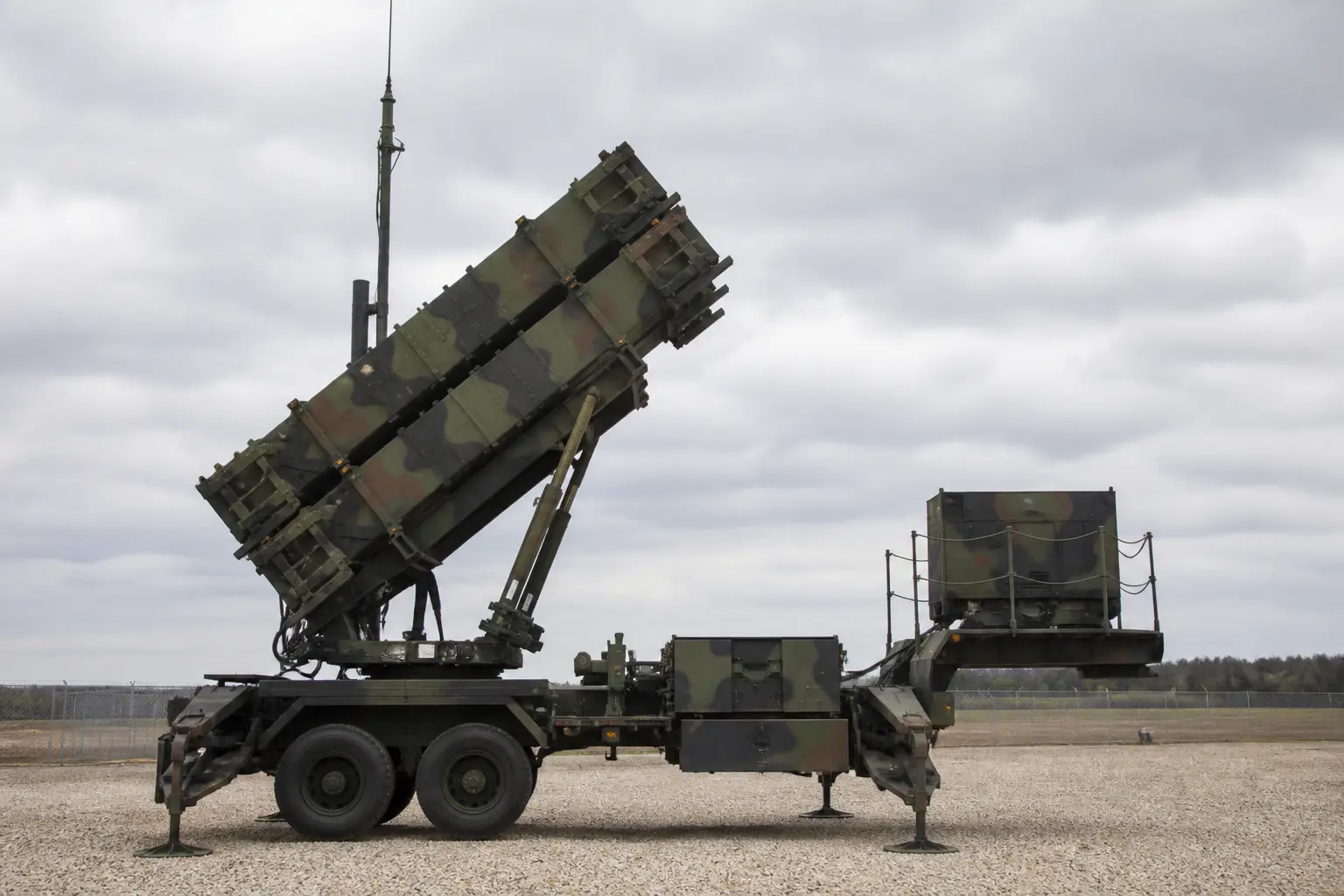 A wide daylight shot of a Patriot missile-defense launcher with its raised canister tubes deployed on a gravel battery site under an overcast sky.