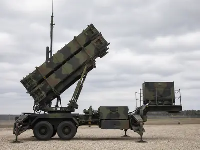 A wide daylight shot of a Patriot missile-defense launcher with its raised canister tubes deployed on a gravel battery site under an overcast sky.