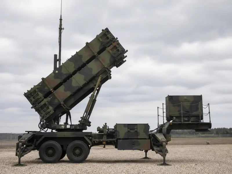 A wide daylight shot of a Patriot missile-defense launcher with its raised canister tubes deployed on a gravel battery site under an overcast sky.
