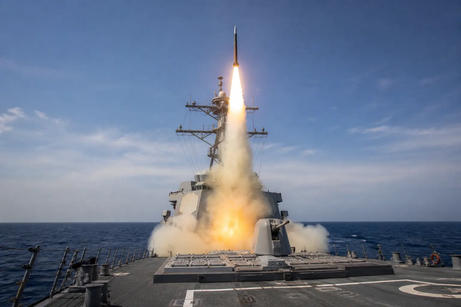 A wide daytime shot of an SM-3 interceptor missile blasting upward from the vertical launch cells on the deck of a US Navy Aegis destroyer at sea, trailing exha
