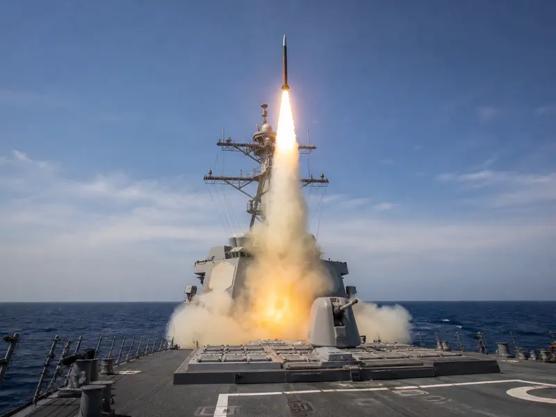A wide daytime shot of an SM-3 interceptor missile blasting upward from the vertical launch cells on the deck of a US Navy Aegis destroyer at sea, trailing exha