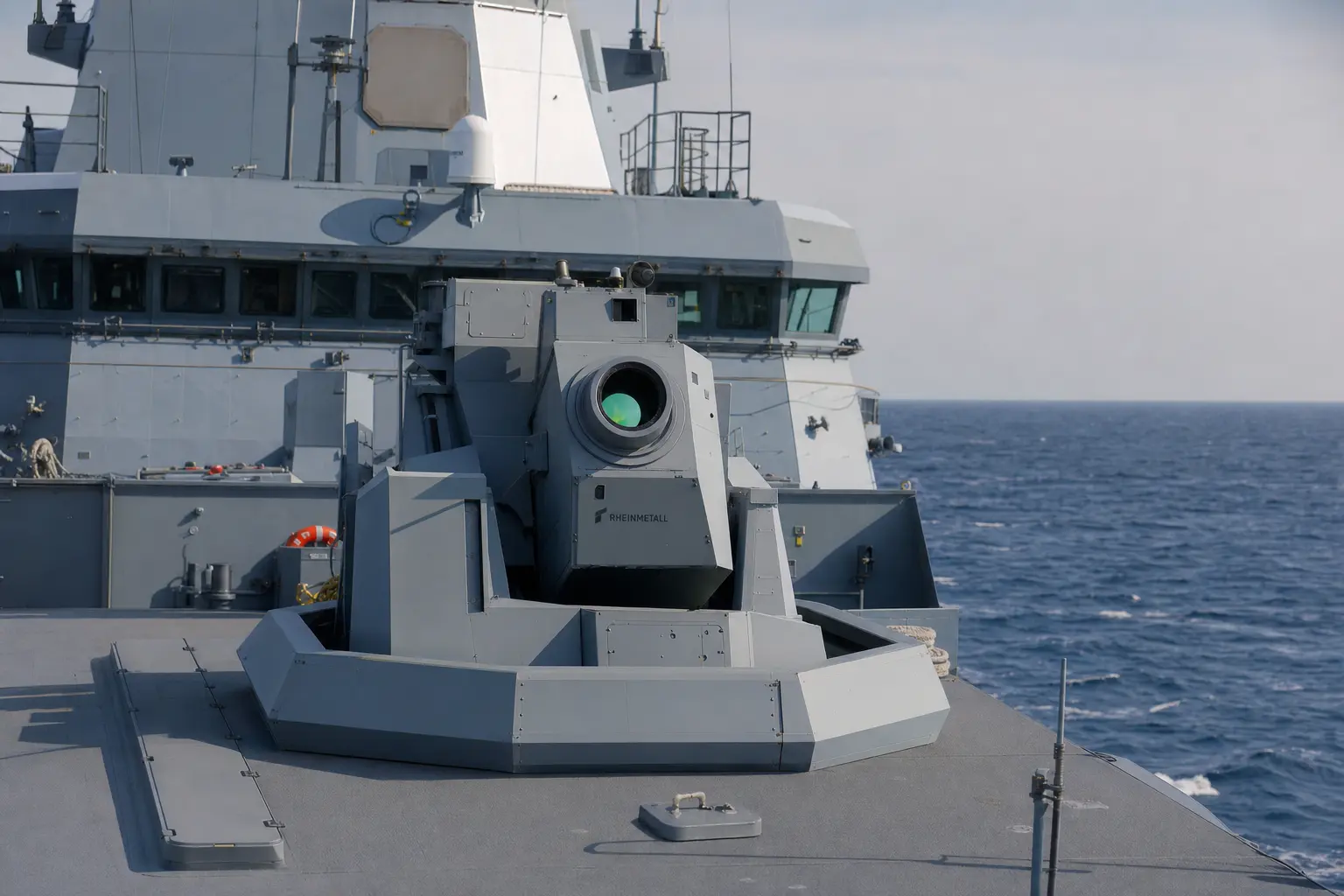 A wide daytime deck-level shot of a German Navy frigate at sea with a high-energy laser weapon demonstrator turret mounted on its deck, Rheinmetall lettering vi