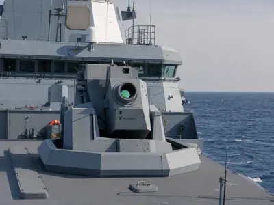 A wide daytime deck-level shot of a German Navy frigate at sea with a high-energy laser weapon demonstrator turret mounted on its deck, Rheinmetall lettering vi