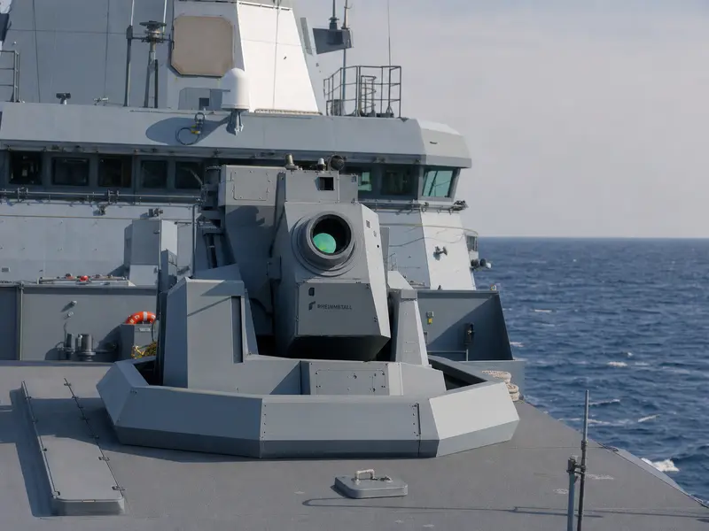 A wide daytime deck-level shot of a German Navy frigate at sea with a high-energy laser weapon demonstrator turret mounted on its deck, Rheinmetall lettering vi