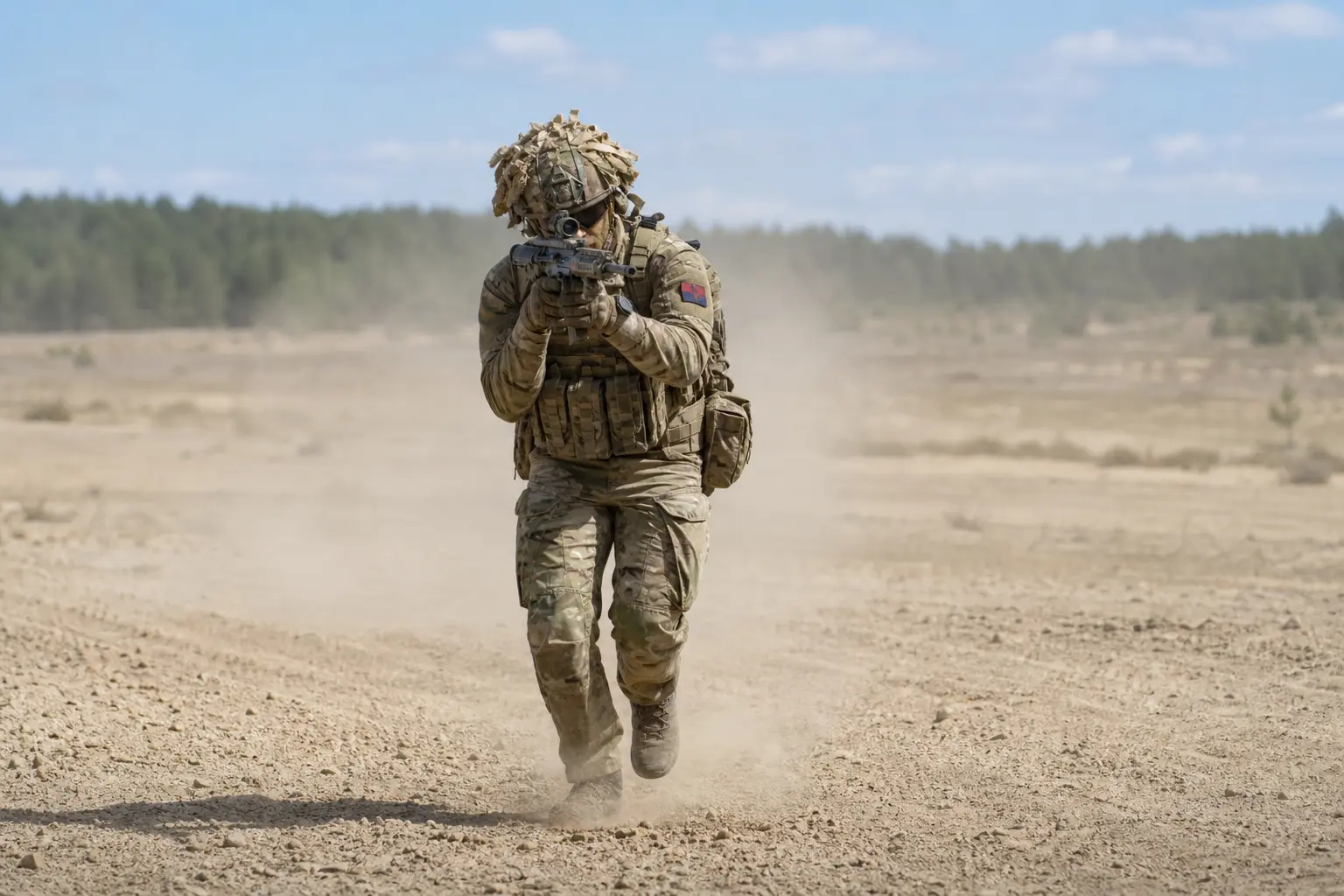 A wide daylight shot of British Army soldiers in camouflage moving across an open training range during a live exercise, weapons raised and dust rising behind t