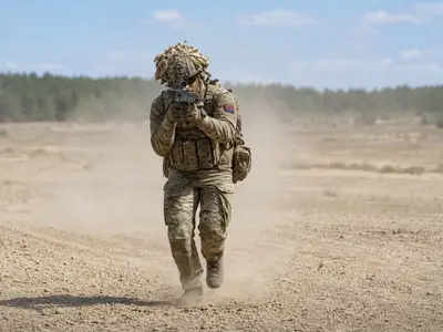 A wide daylight shot of British Army soldiers in camouflage moving across an open training range during a live exercise, weapons raised and dust rising behind t