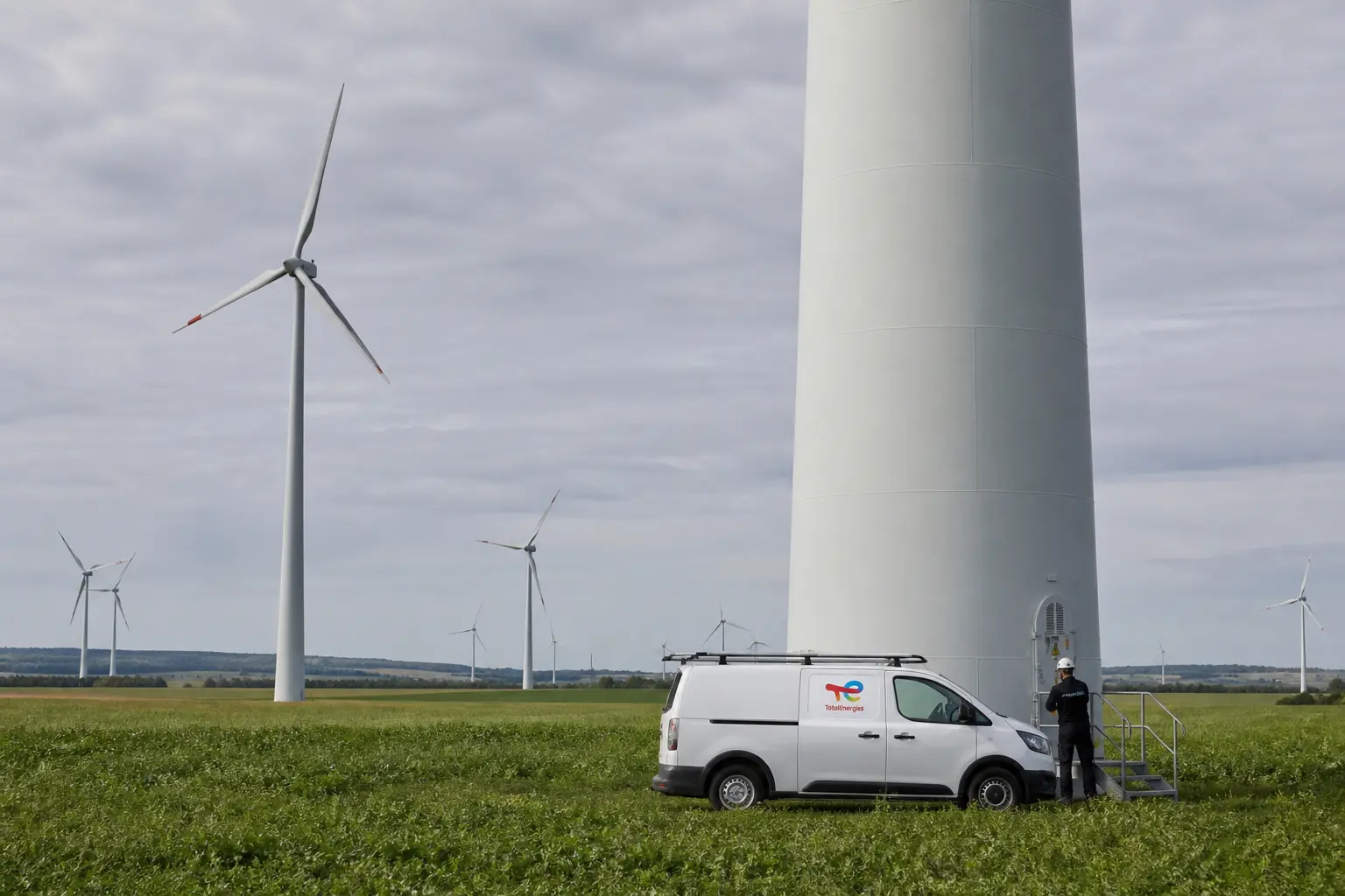 A wide daytime establishing shot of a European onshore wind farm, rows of white turbines turning across green farmland under an overcast sky, with a maintenance