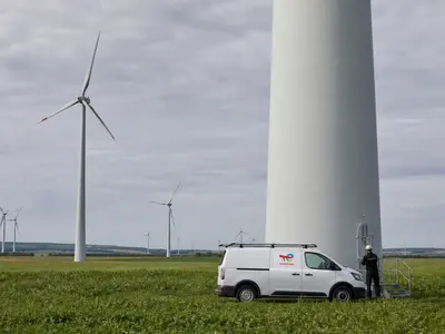 A wide daytime establishing shot of a European onshore wind farm, rows of white turbines turning across green farmland under an overcast sky, with a maintenance