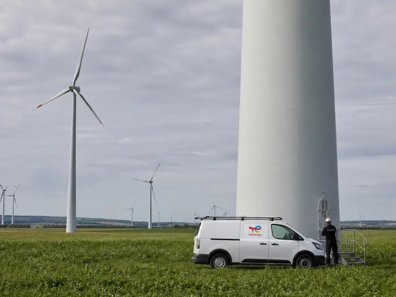 A wide daytime establishing shot of a European onshore wind farm, rows of white turbines turning across green farmland under an overcast sky, with a maintenance