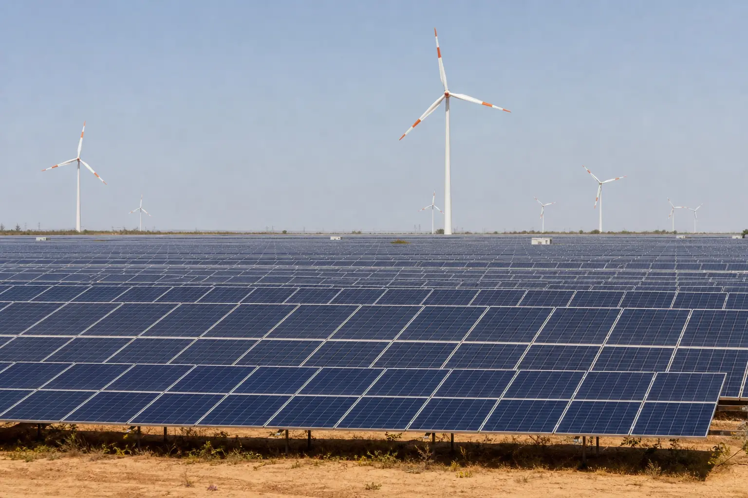 A wide establishing shot of a large operating utility-scale solar farm in India under bright daylight, rows of photovoltaic panels stretching across arid open l