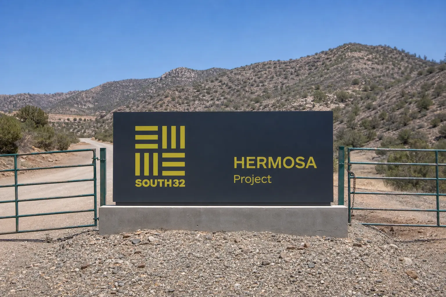 A straight-on daylight view of the South32 company sign at the entrance gate to the Hermosa project site, with graded access road and scrubland ridges of the Co