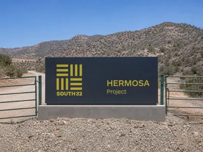 A straight-on daylight view of the South32 company sign at the entrance gate to the Hermosa project site, with graded access road and scrubland ridges of the Co